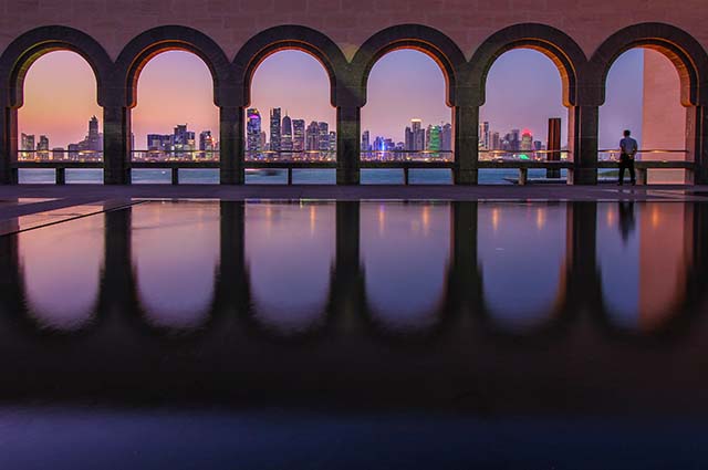 Old stone arches show a restricted view of the Doha skyline at sunset with the multiple skyscrapers lit up with a orange hue and their lights flickering. In front is the water front splitting the city from the ancient arches