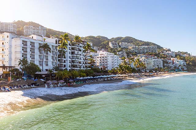 Beach front of Puerto Vallarta with the waves crashing agains the sandy beach, which is lined with sun loungers. Behind the buildings rise up and climb with the rolling lush green hills