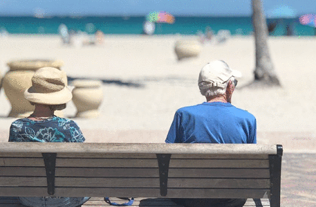 an older couple sat on a beach by the seafront, with sand and blue sky